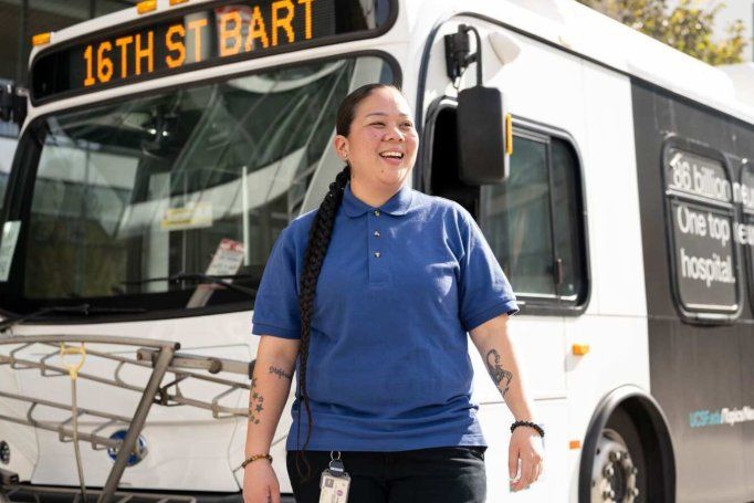 Shuttle driver stands in front of bus smiling