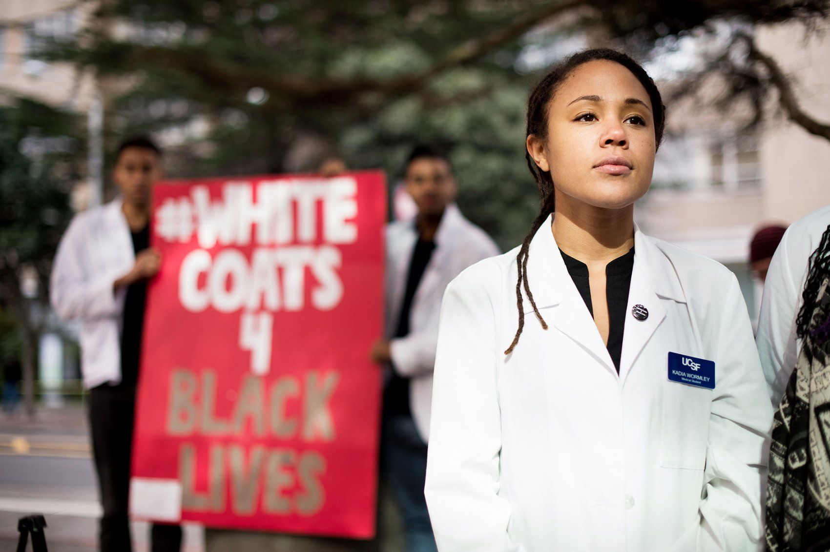 a UCSF student stands in front of a sign that says White Coats 4 Black Lives
