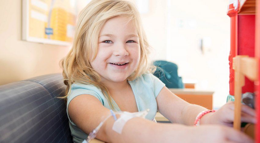 Child smiling in a hospital chair, with a medical line in her arm.