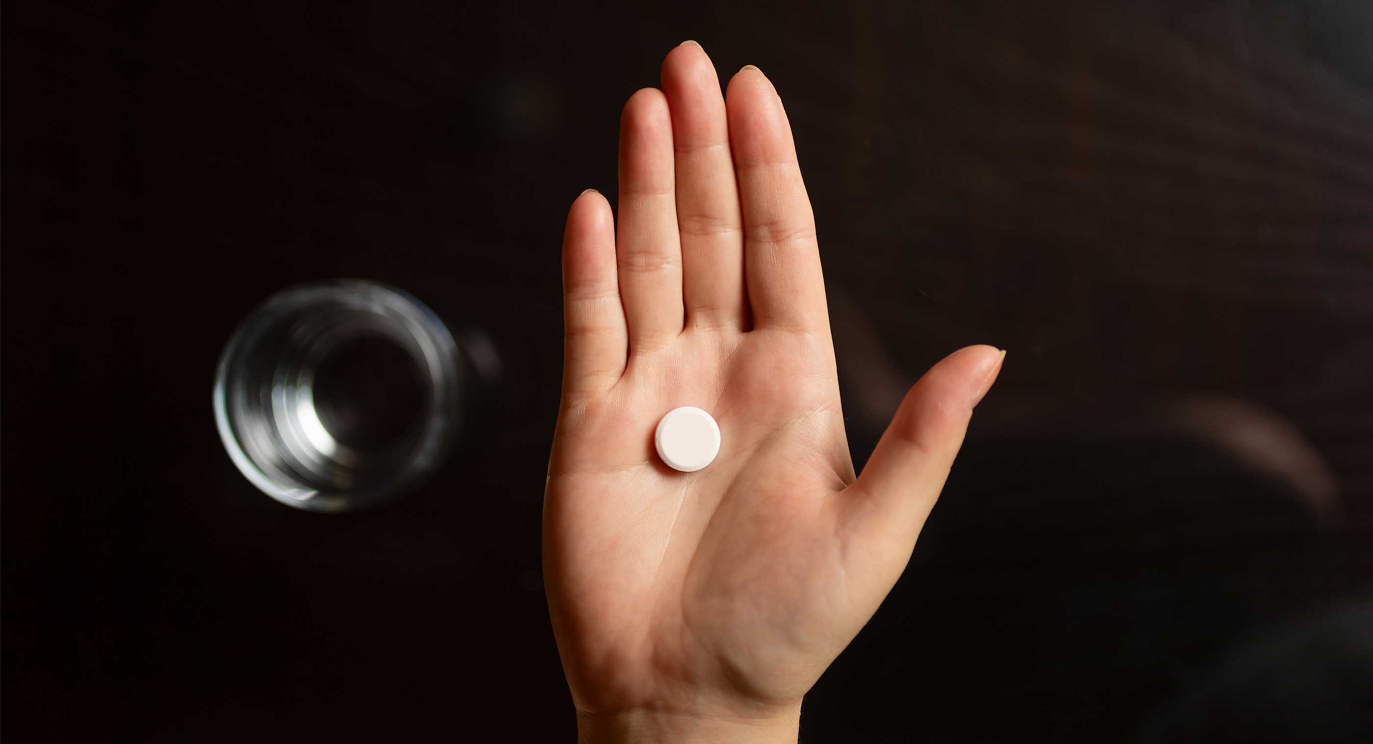 A woman's hand holds a single pill. On a table below the hand is a glass of water.