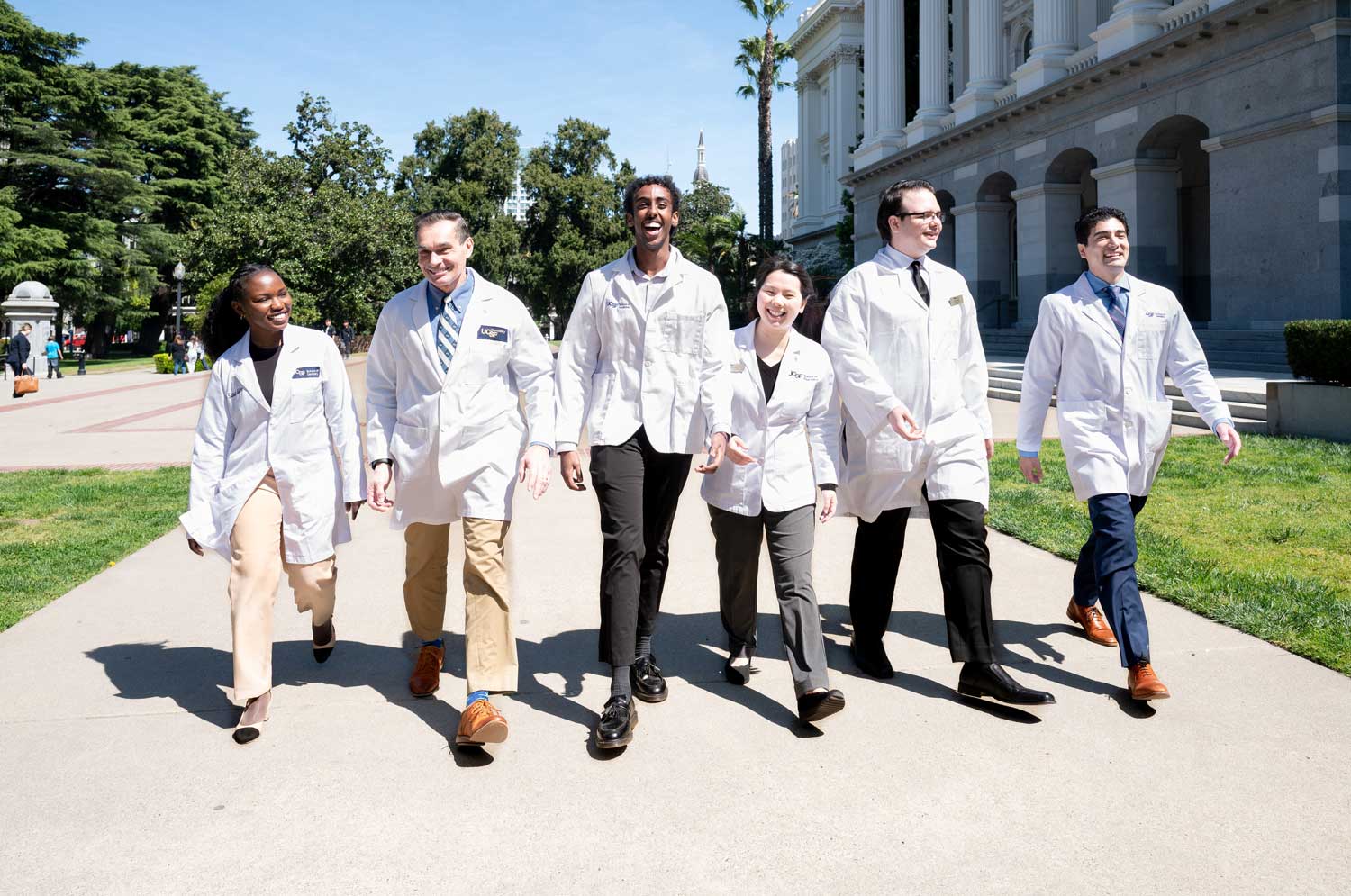 A group of medicine, pharmacy, and dental students wearing white coats smile as they walk together on a sunny pavement outisde the California State Capitol building in Sacramento.