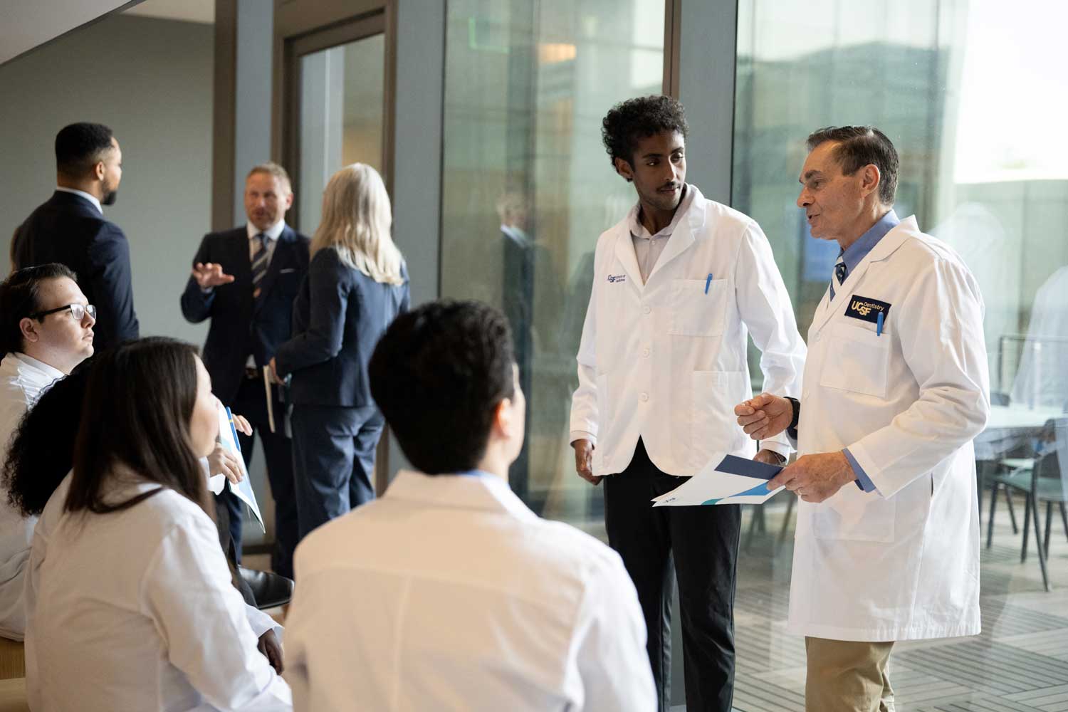 A group of medical, dental, and pharmaceutical students wearing white coats sit in a glassy atrium.