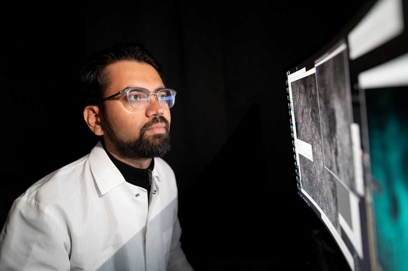 Vijay Namboodari sits in a dark room and looks at scientific images on two desktop computers.