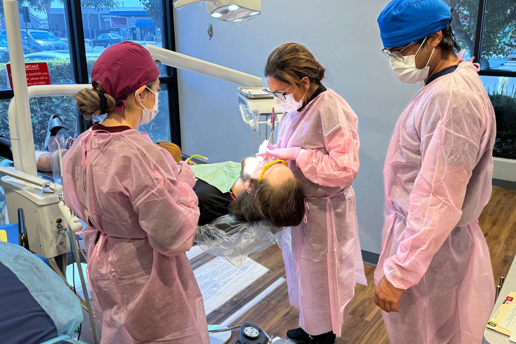 three students look over client in dental office chair with light over mouth.