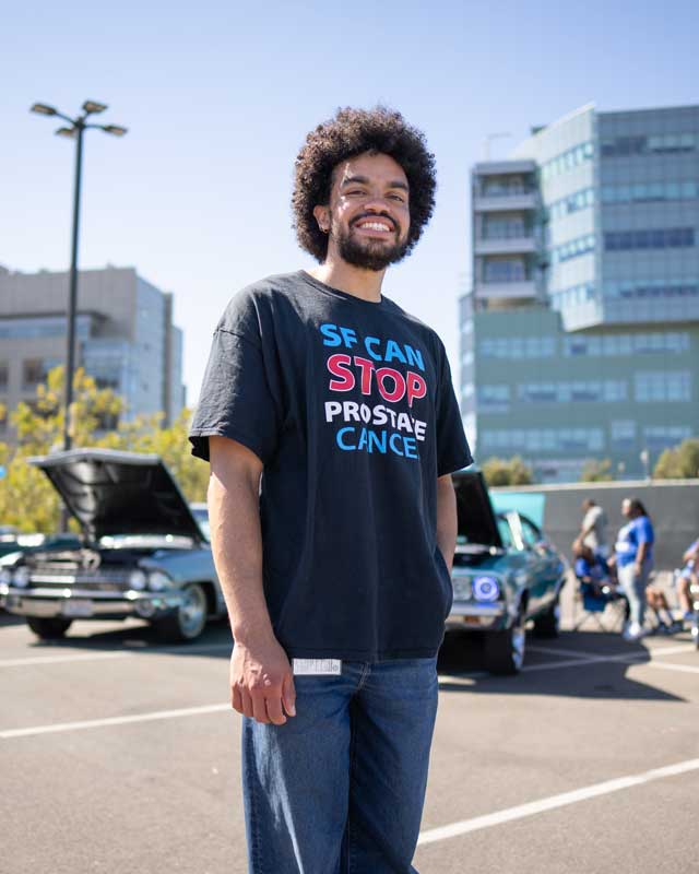 A young Black man named Joseph Egbunikeokye smiles as he stands in front of vintage cars at Mission Bay. He wears a T-shirt that reads "S F CAN STOP PROSTATE CANCER."