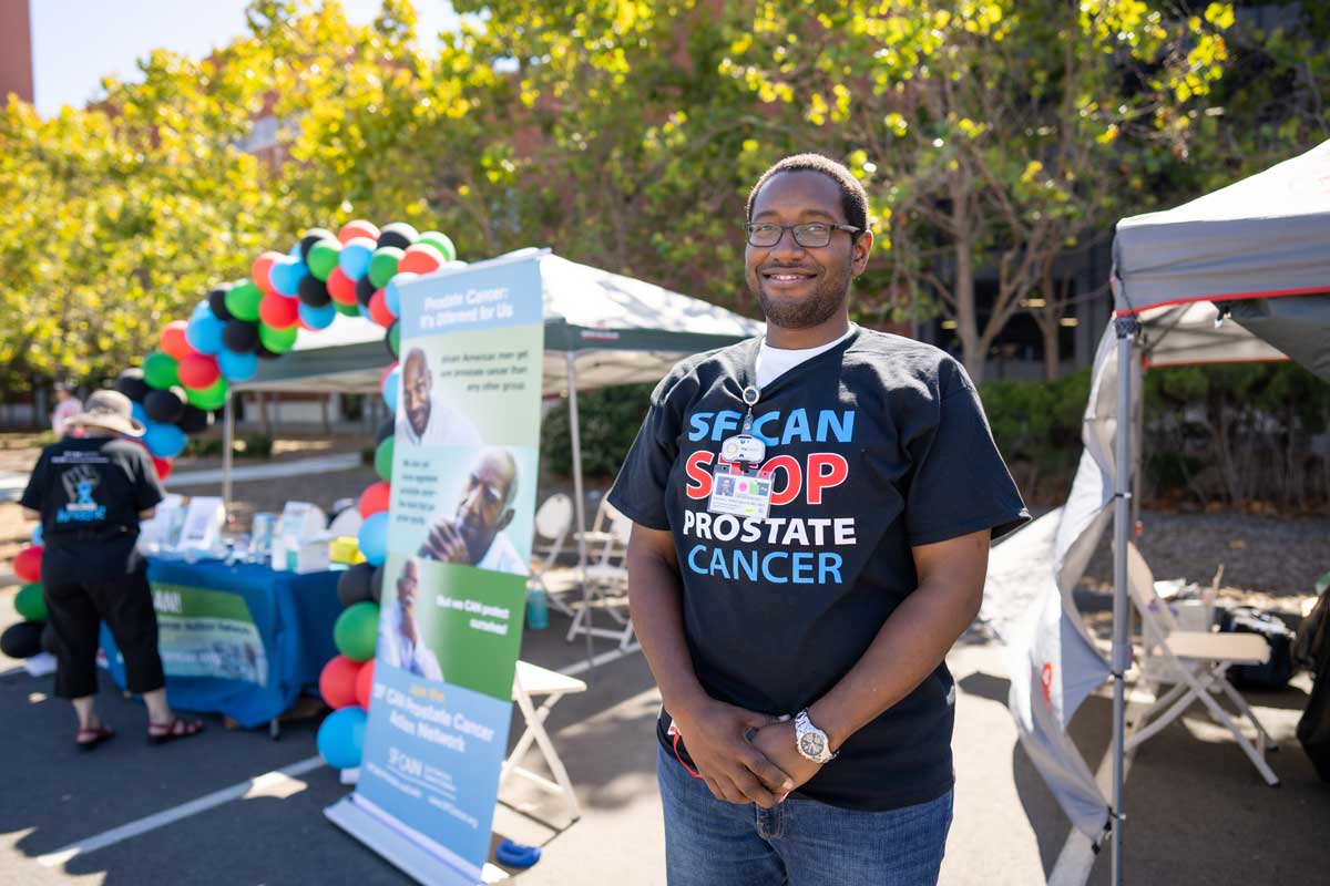 A young African American man named Samuel L. Washington poses in front of an informational booth. He wears a T-shirt that reads "S F CAN STOP PROSTATE CANCER."