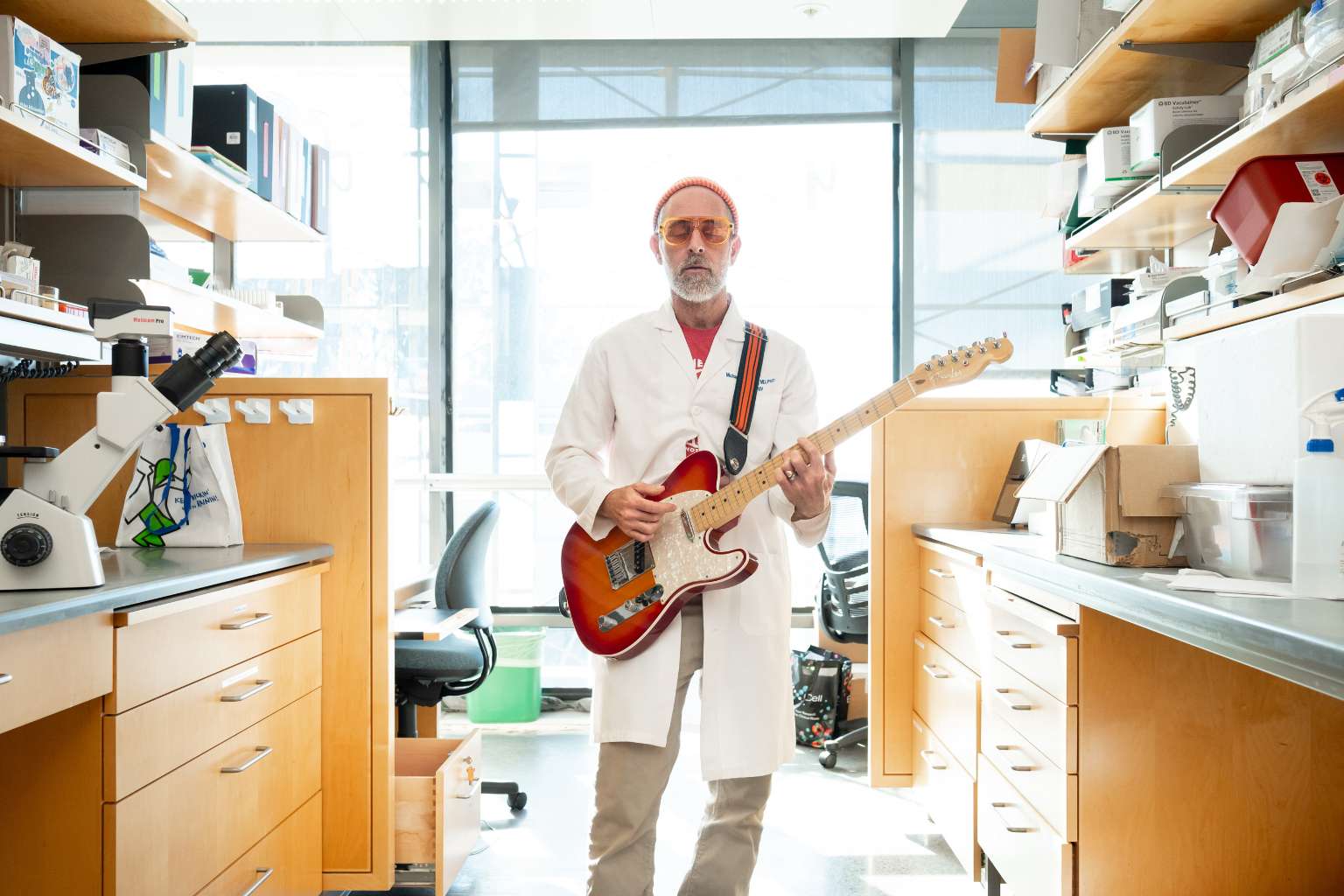 Man in beanie and lab coat plays electric guitar.