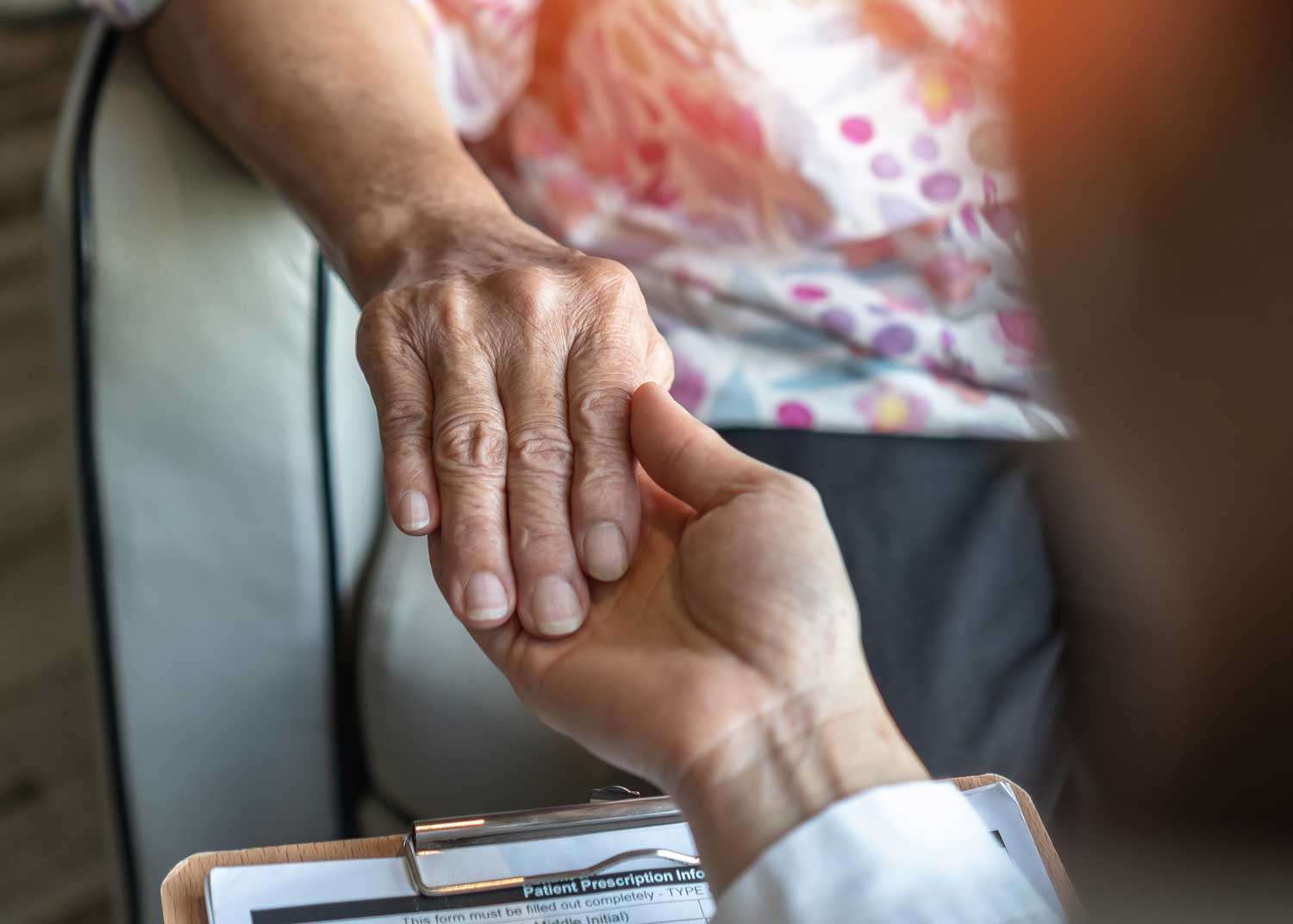 A caregiver holds an elderly woman's hand in comfort.