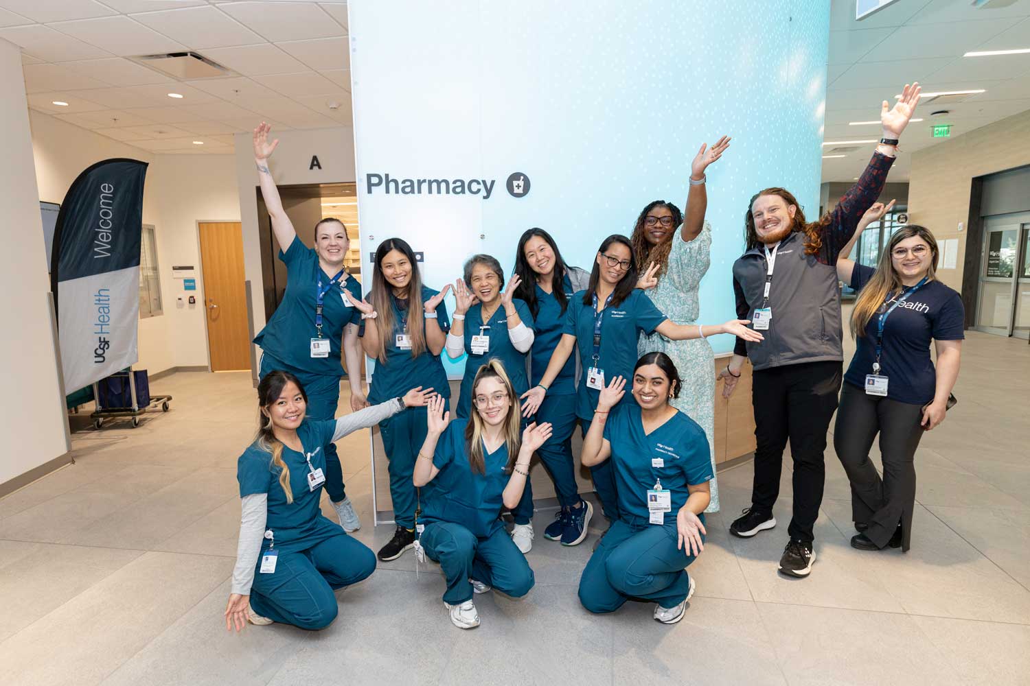 A group of pharmacy technicians, pharmacists, and other staff pose happily at the entrance of a new Pharmacy at UCSF Mission Bay.