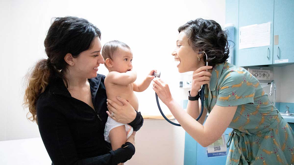 A doctor smiles as she holds up a stethoscope to an infant held in its mother's arms.