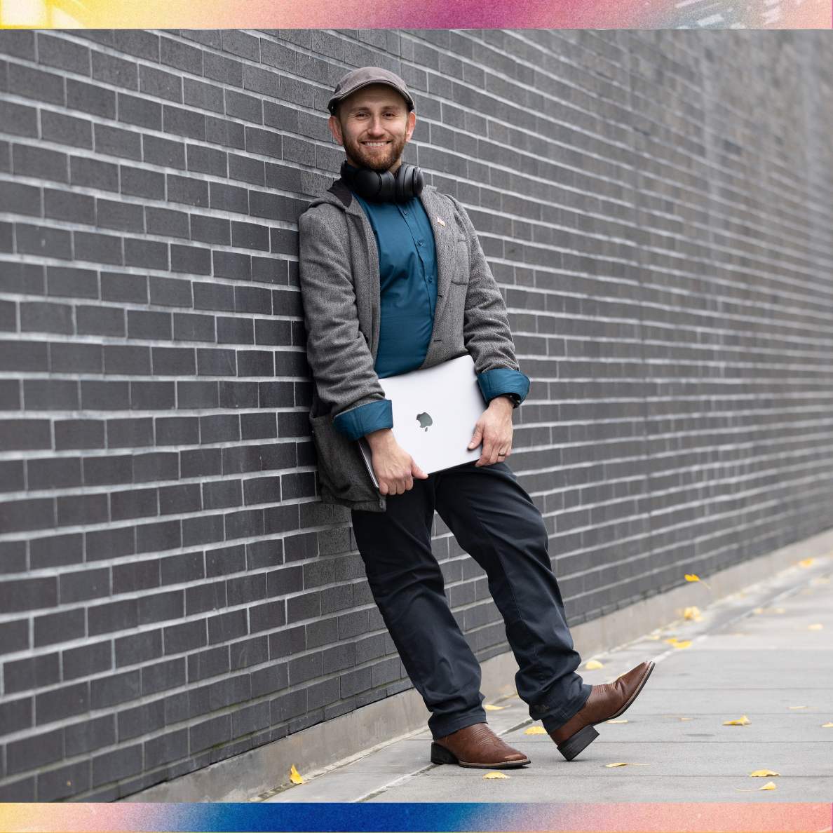 Jonathan stands against brick wall holding laptop.