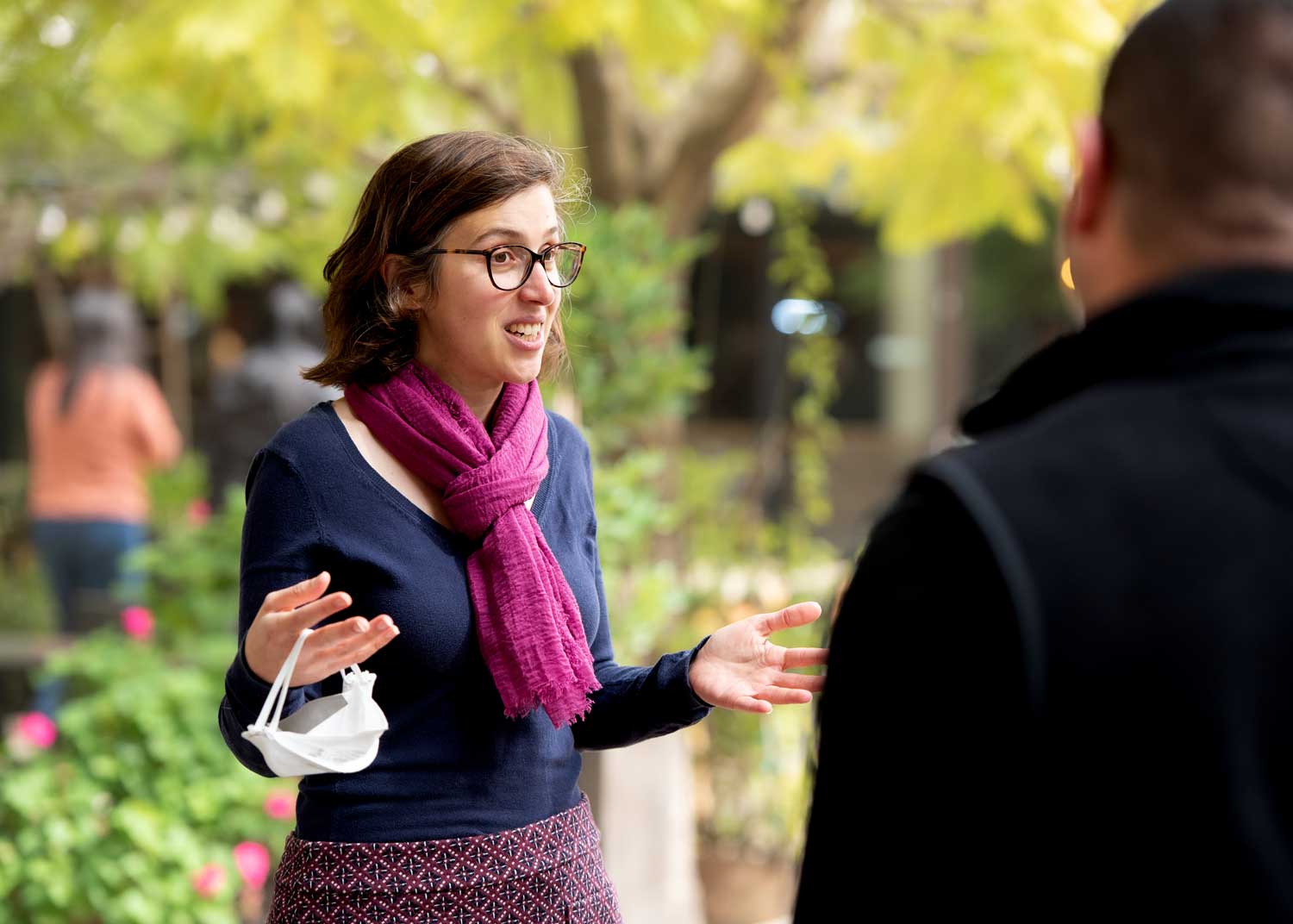 Alison Cohen holds a protective facial mask while speaking with a colleague outdoors.