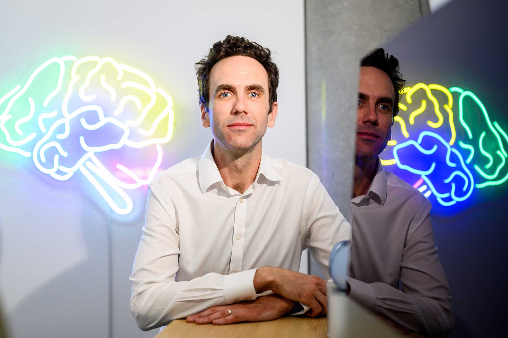 Simon Little sits in his Mission Bay lab in front of a neon sign in the figure of a brain, which is also reflected on his computer screen.