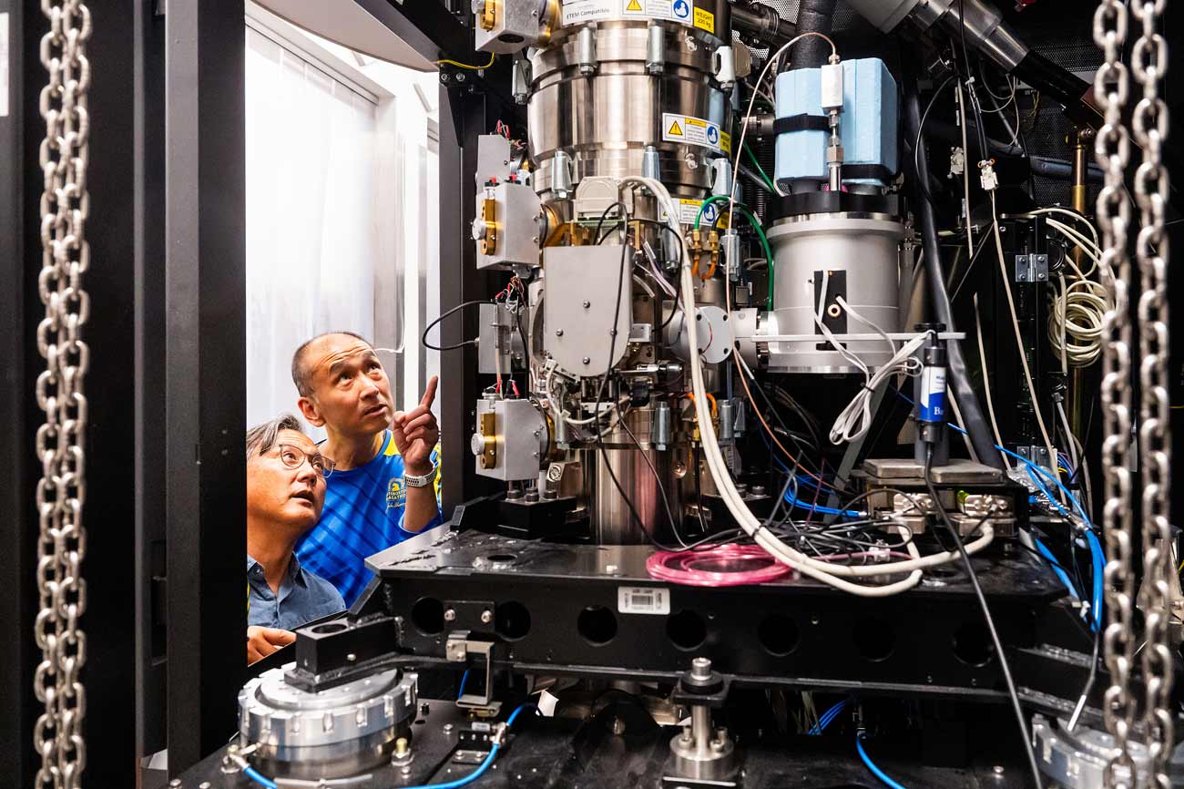 Stephen Nishimura and Yifan Cheng look up at a large electron microscope.