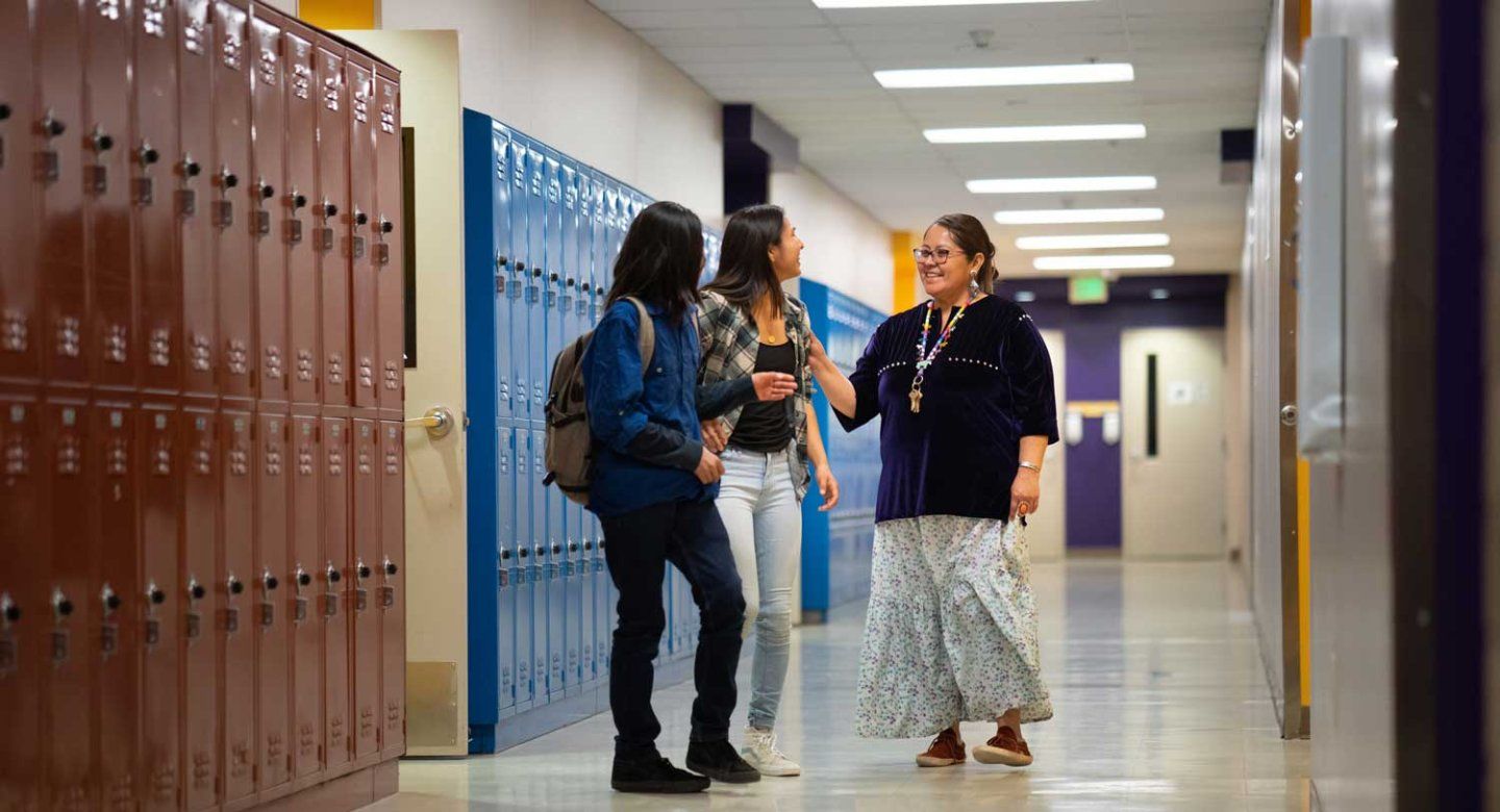 A teacher laughs with two female high school students at a school hallway.