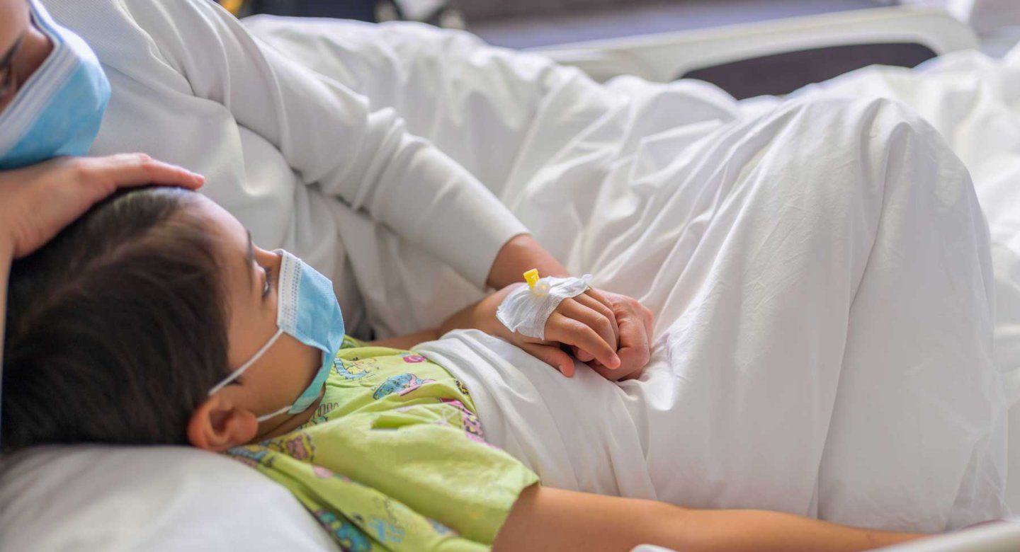 A child and his parent lie together on a hospital bed. They both wear surgial masks.