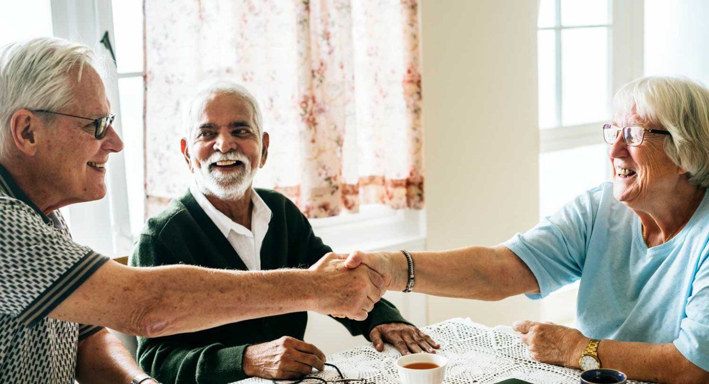 A group of three elerdly people smile together as they sit at a sun-lit kitchen.