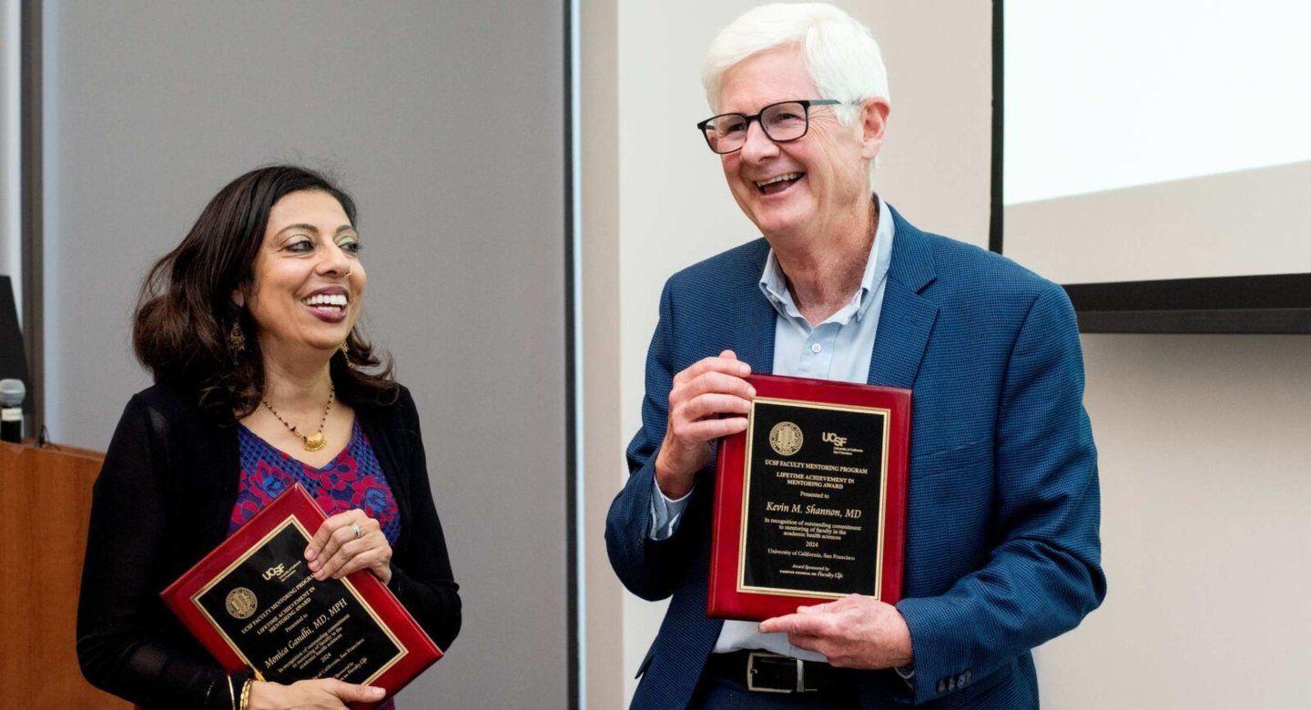 Two recipients hold awards and smile at each other. 