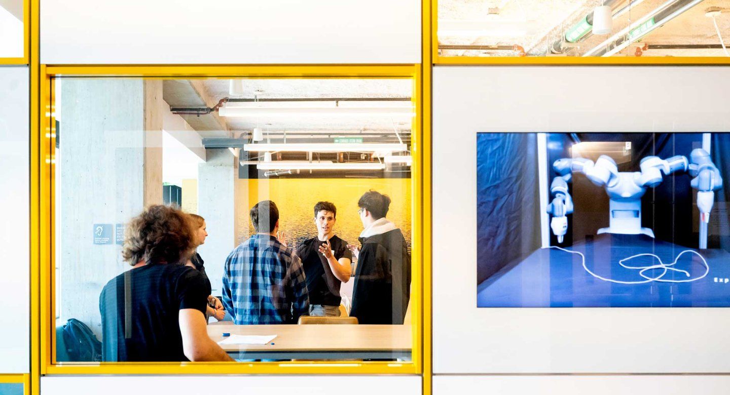 A perspective from outside a glassy classroom window shows students speaking with a professor. A screen on the glassy wall shows a robot doing precise movements.