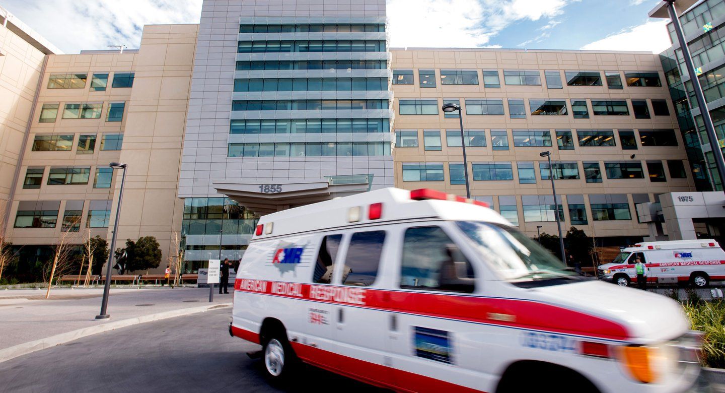 A blurred emergency vehicle speeds out of a hospital parking lot.