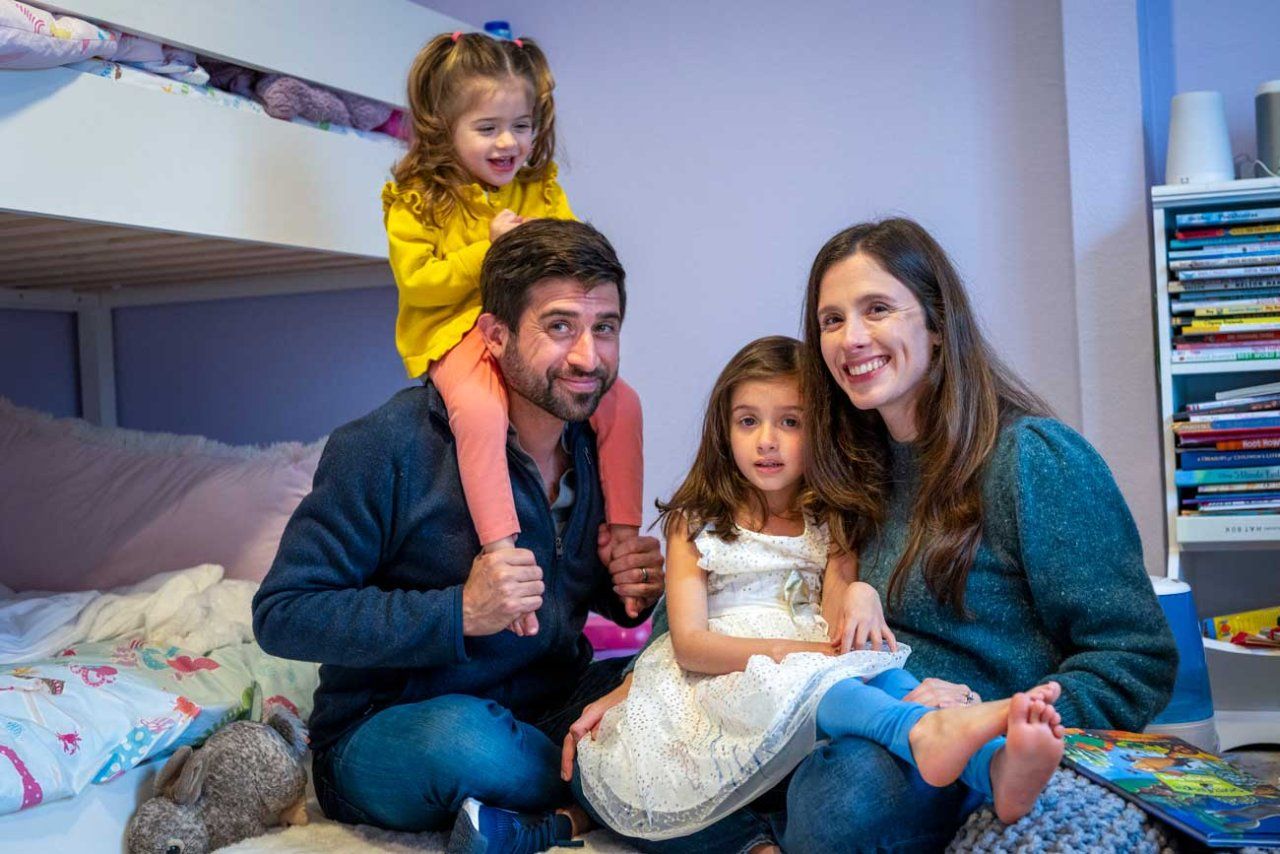 Two-year-old Emiliana laughs as she shits on her father's shoulders. Her father, mother, and younger sister smile as they sit on the girls' bedroom.