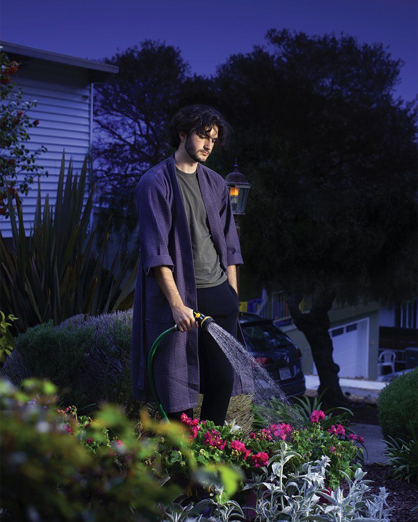 Photo of a young man in a bath robe, watering plants in the yard in the middle of the night.