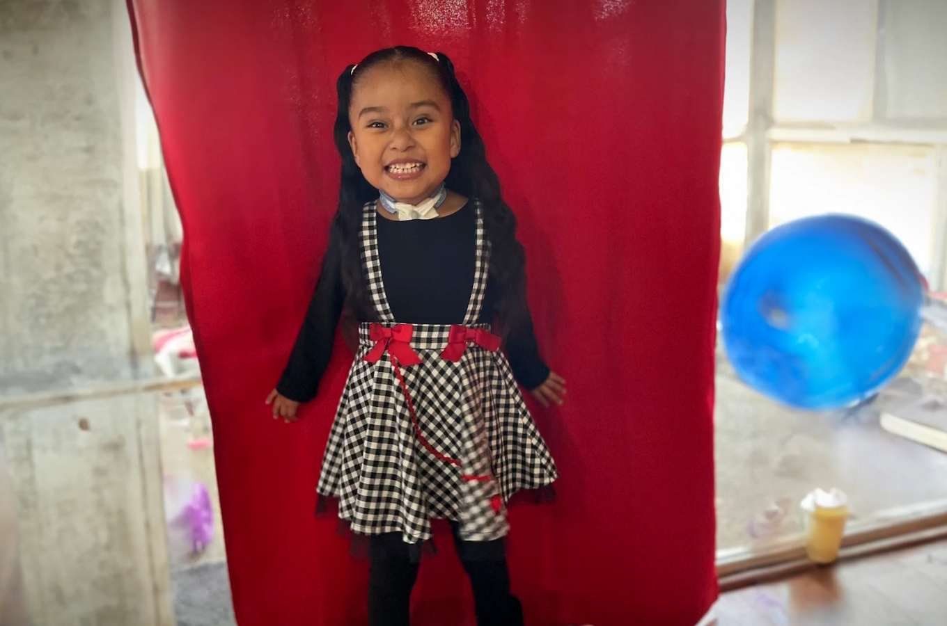 Little girl poses in black dress against red curtain.