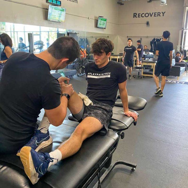 A young athlete named Joey Papa sits on a physical therapy bench at a gym while a medical professional treats his knee with a massage.