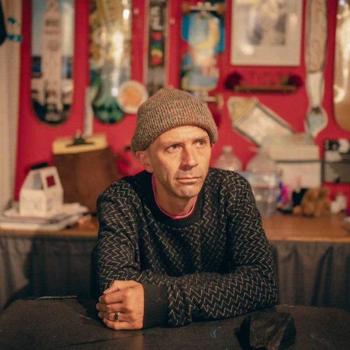 Former pro skateboarder Shawn Connolly, a Caucasian man wearing a beanie, sits contemplatively in front of a wall decorated with skateboards.