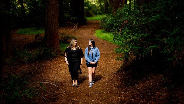 A mother and daugther walk pensiveley amongst a dark backdrop of redwood trees.
