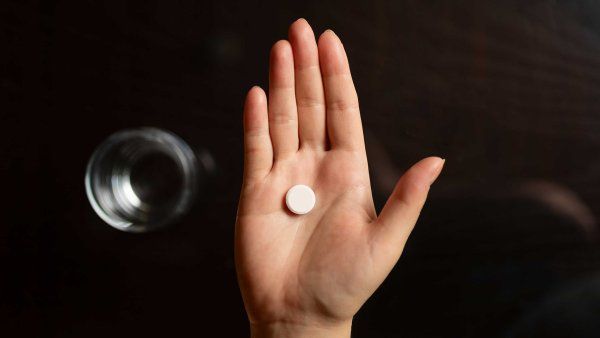 A woman's hand holds a single pill. On a table below the hand is a glass of water.