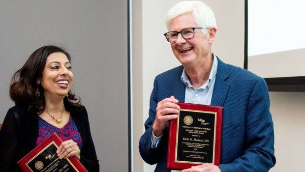 Two smiling individuals hold awards and smile at each other.