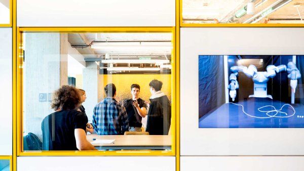 A perspective from outside a glassy classroom window shows students speaking with a professor. A screen on the glassy wall shows a robot doing precise movements.