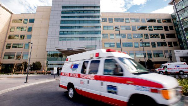 A blurred emergency vehicle speeds out of a hospital parking lot.