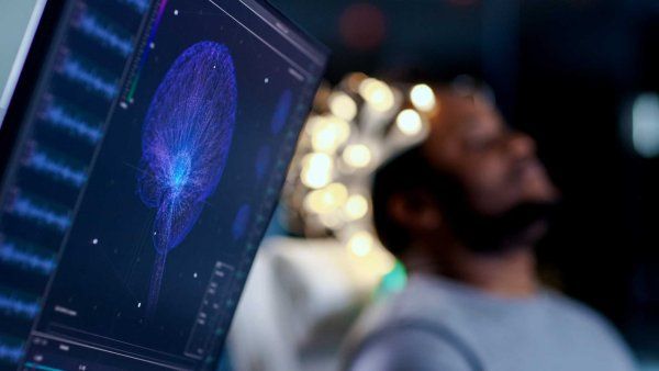 Computer monitors showing E E G reading on a graphical brain model; in the background, a man wears a brainwave reading headset while he sits with his eyes closed.