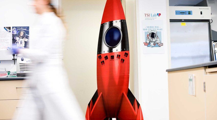 A large cutout of a red space rocket stands in the UCSF lab of Sonja Schrepfer and Tobias Deuse. A lab assistant in a white lab coat walks by.