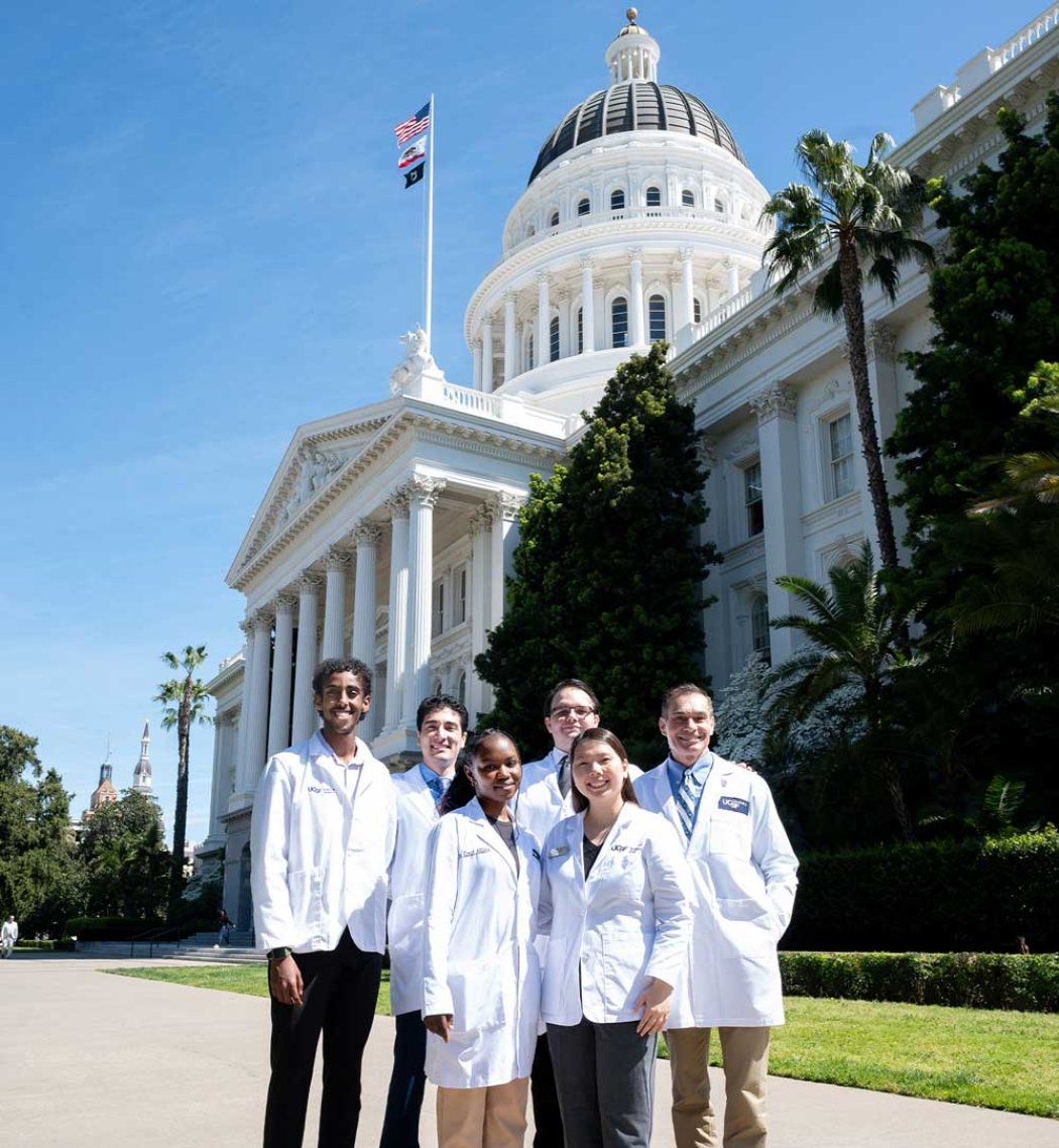 A group of medicine, pharmacy, and dental students, and a faculty member, all wearing white coats, smile as they stand on a sunny pavement outisde the California State Capitol building in Sacramento.
