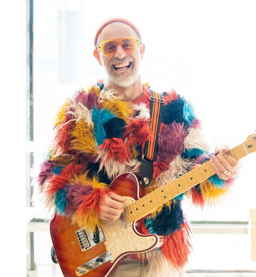 Man in fluffy coat and beanie plays electric guitar in laboratory.