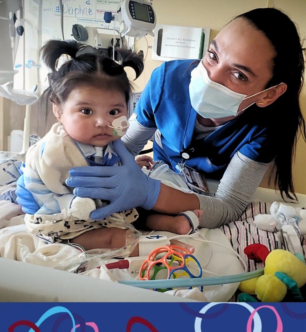 Baby surrounded by toys in hospital bed cared for by nurse.