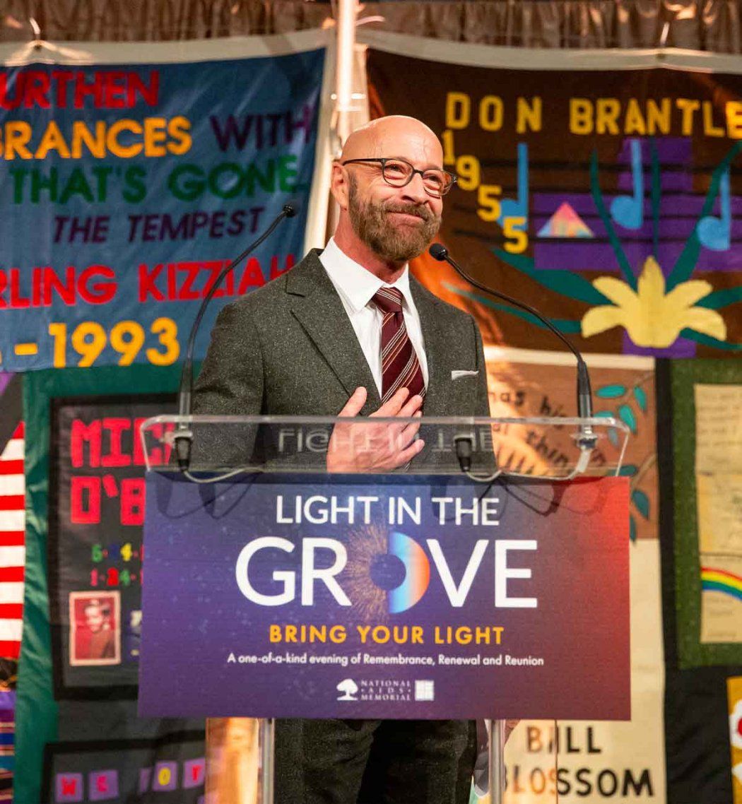 Dan Bernal smiles as he stands at a podium that has a sign that reads "Light in the Grove." In the background are multicolored flags with names of previous award winners.