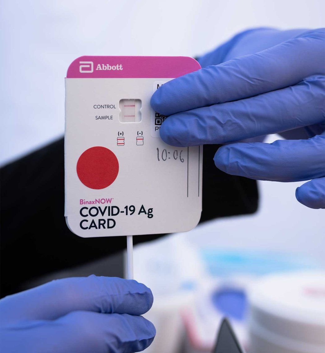 Two gloved hands hold a rapid antigen test card showing a positive COVID-19 result.